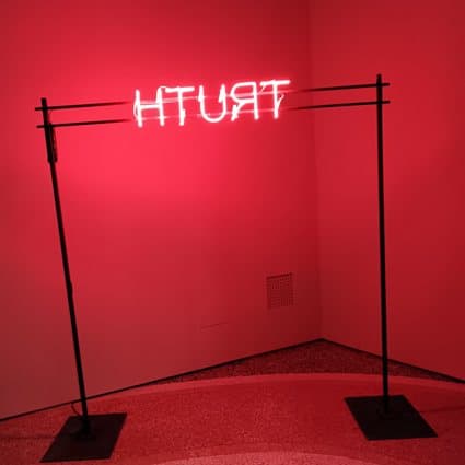 Scritta al neon rossa TRUTH rovesciata, sospesa tra due aste in una stanza rossa
